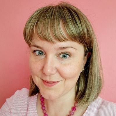 Angharad is smiling against a mid-pink background. She is wearing a light pink top, red bead necklace and pearlescent earrings. She has shoulder length light brown and blonde hair, and is wearing natural looking makeup. 