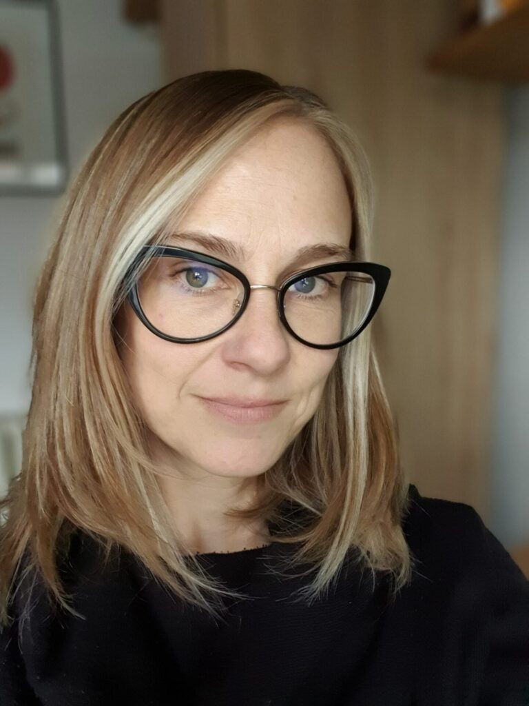 Cat is smiling stood against a light background. She is wearing large dark framed glasses and a black top. She has light brown and blonde hair which is shoulder length and falls slightly over her glasses. 