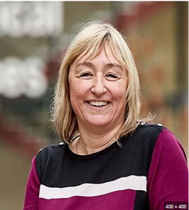 Karen is smiling stood against a blurred outdoors background. She is wearing a black and magenta jumper. She has shoulder length blonde hair with a fringe. 