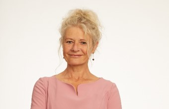 Symone is smiling against a white background. Her blonde hair is worn in a messy up-do. She is wearing a blush dress and hanging jewelled earrings. 