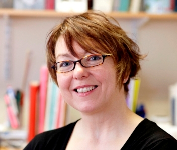 Marion is stood smiling against a light office background. She is wearing a black top and small rectangular shaped glasses. She has short red brown hair in a pixie style cut. 
